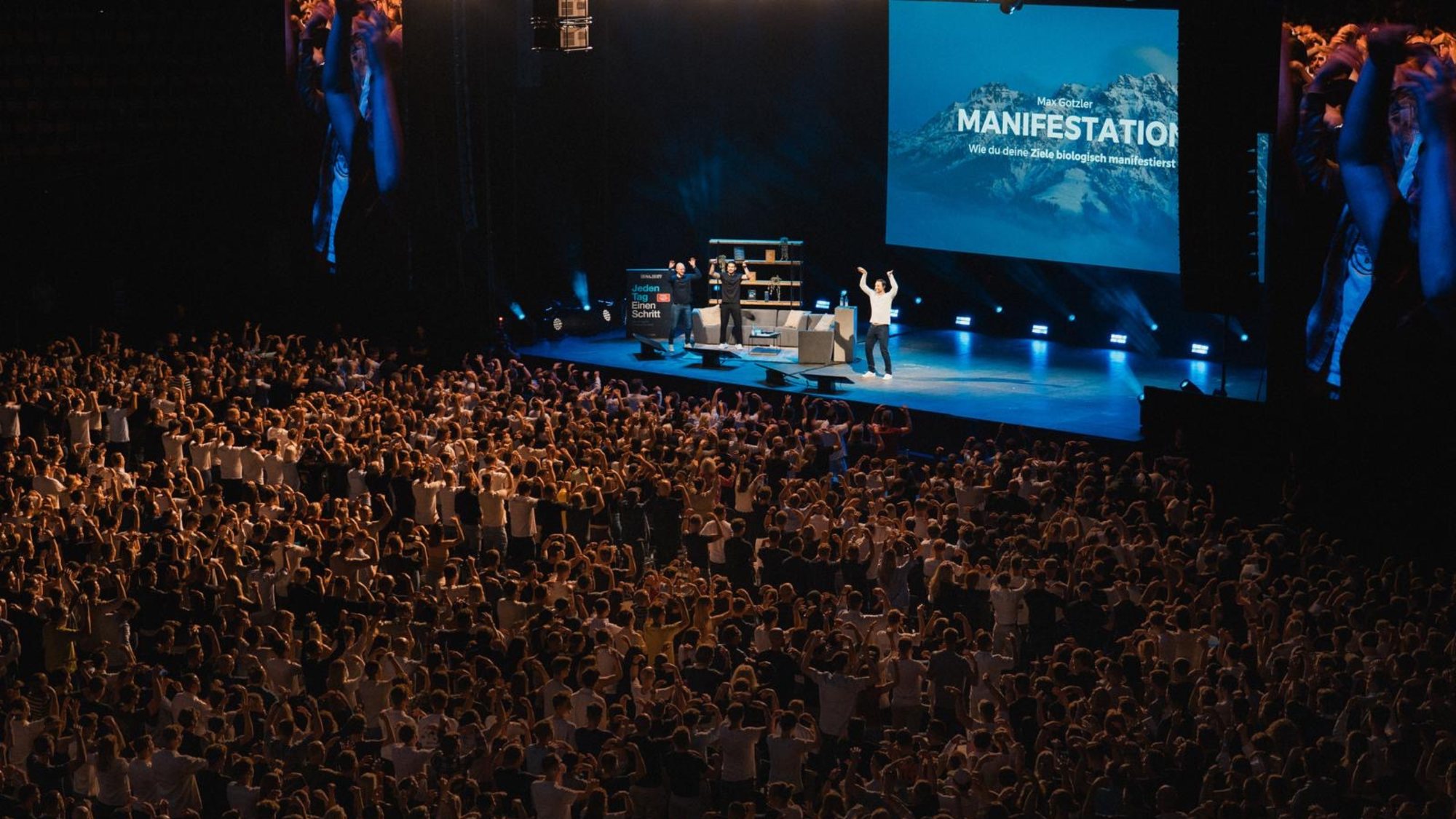 Max Gotzler auf einer großen Bühne vor mehreren tausend Menschen, auf der Leinwand: „Manifestation“